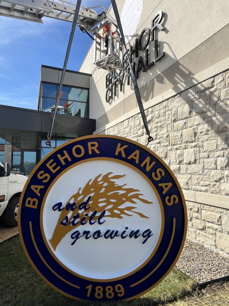 Sign installation at Basehor City Hall in Kansas: a circular Basehor, Kansas seal sign with a gold wheat emblem and the words "and still growing, 1889" hangs from lifting straps under an aerial lift, in front of the building's stone facade and dimensional letters.
