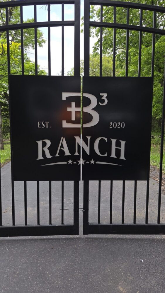 Black double gate with a large black sign reading 'B3 Ranch' and 'EST. 2020'; gate bars visible with greenery beyond.