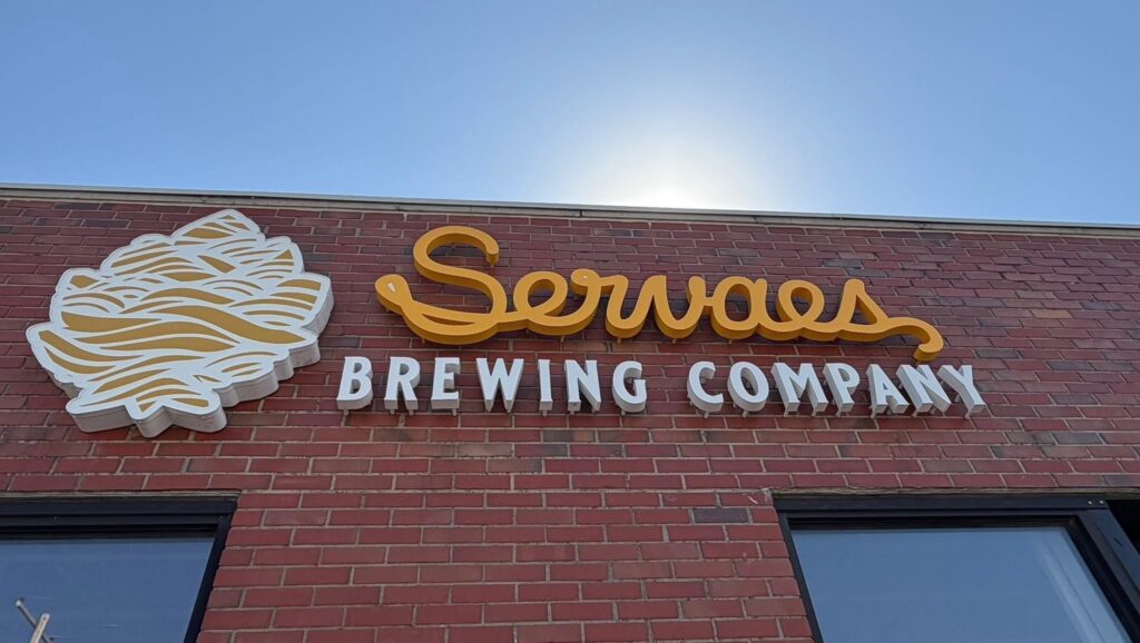 IMG_6019 » Prairie Fire Signs & Graphics Modern sign design displayed on a storefront featuring bold colors and clear branding to attract customer attention and influence purchasing decisions. Sign on a brick wall reading'Sevens Brewing Company' with a yellow cursive logo and a white hop leaf emblem on the left.